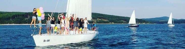 Група млади хора се забавляват на яхта под наем сред живописен воден пейзаж с планински фон и други яхти.